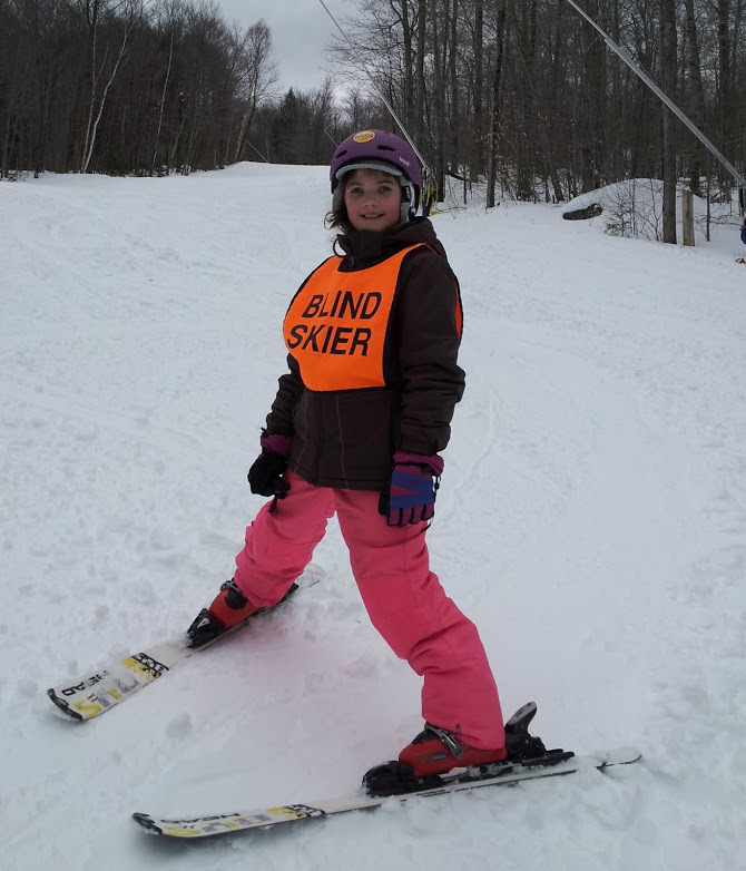 Young Abby on a snowy slope, smiling, wearing winter gear, and an orange ski bib with the words "Blind Skier." Her skis are positioned in a snowplow stance, with her skis angled inward. 