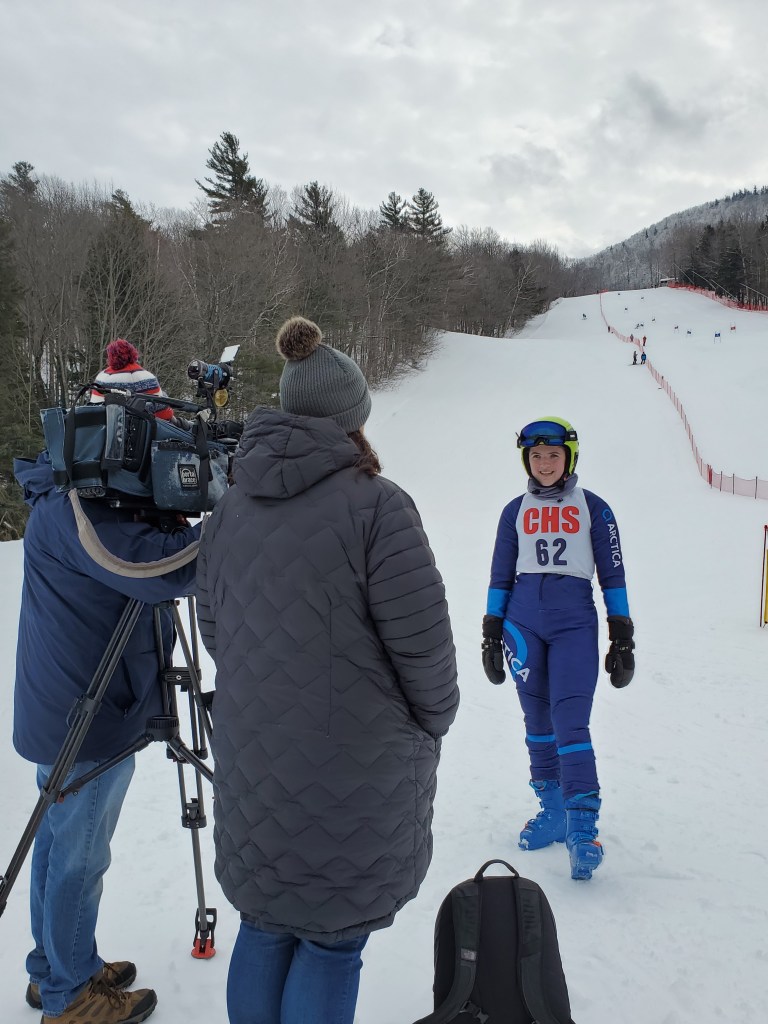 Abby slope side, wearing a blue ski suit and bib that says CHS 82. Video camera is facing her. 
