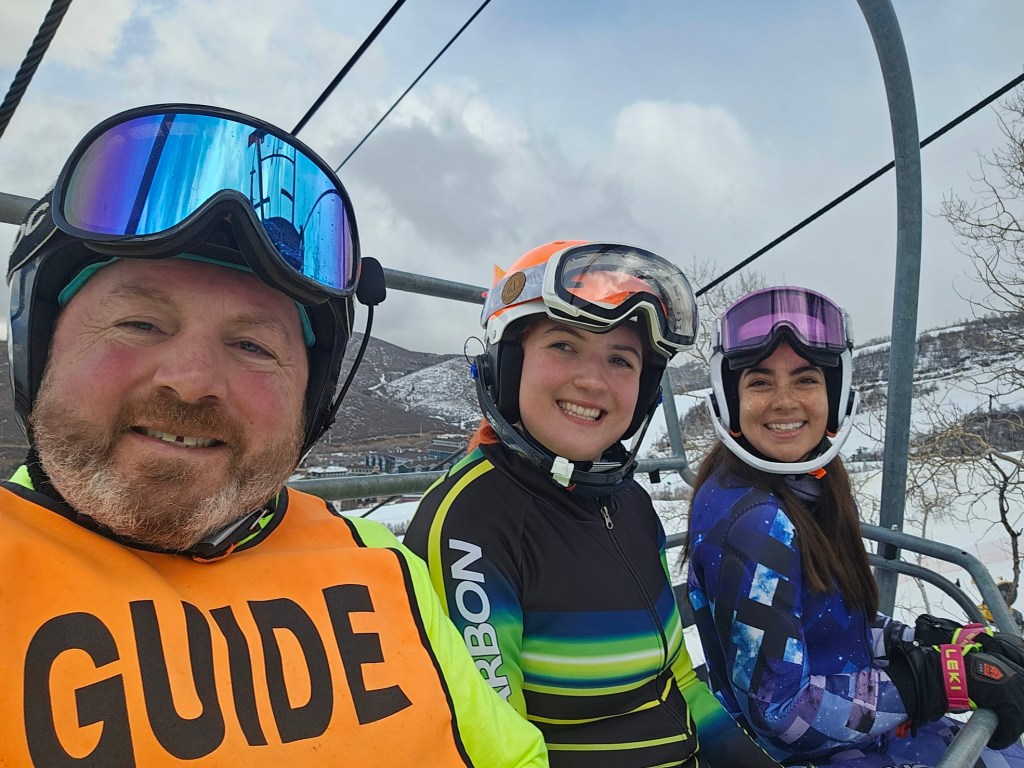 Abby, Chris, and another skiing sitting on a chair lift, smiling. Their goggles are on the top of their helmets. Chris is wearing an orange ski bib that with the words "GUIDE."