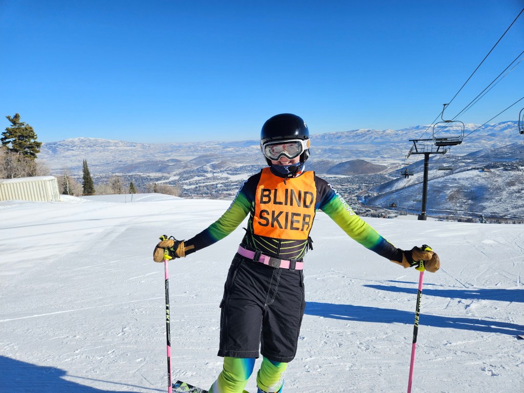Abby smiling, wearing a ski helmet, mask, suit, and an orange "blind skier" bib with a ski pole in each hand on the snow covered slope.