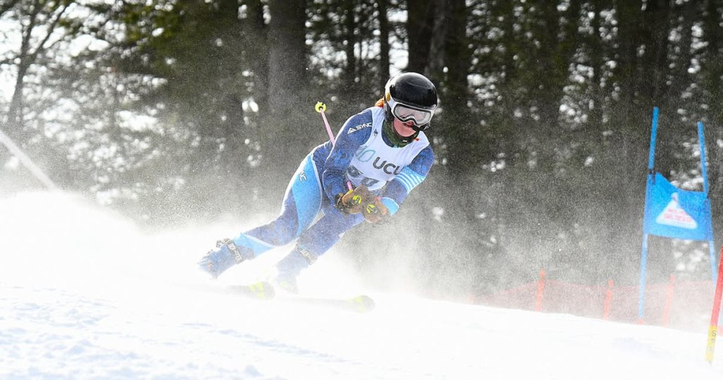 Abby wearing a blue ski suit, helmet, and goggles, downhill skiing. A spray of snow is rising from her skis while making a sharp descent. 