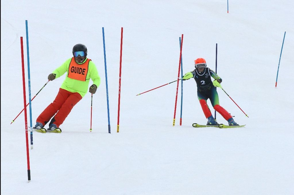 Abby and Chris skiing on a slalom course. On the left, Chris is wearing a bright yellow jacket and orange bib with the words "GUIDE." Abby is skiing on the right, slightly behind her guide, navigating through slalom poles. 
