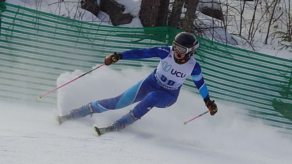 Abby downhill skiing wearing a blue ski suit, a UCU ski bib with the number 50.  Her posture indicating a sharp turn, with left ski angled in the snow, creating a spray of snow behind her. 