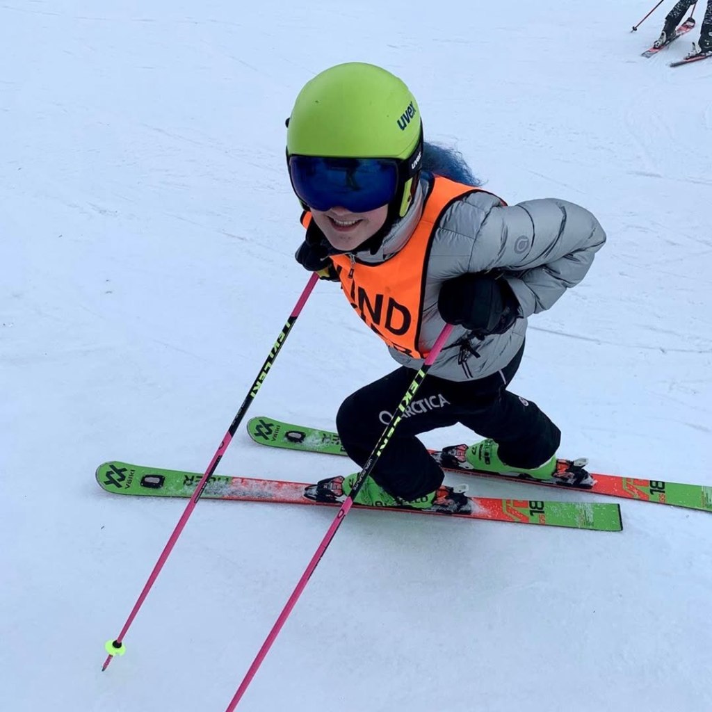 Abby wearing a gray coat, yellow helmet, orange ski bib and ski goggles, smiling while looking upward. She is wearing skis and holding a ski pole in each hand. on a ski slope. 