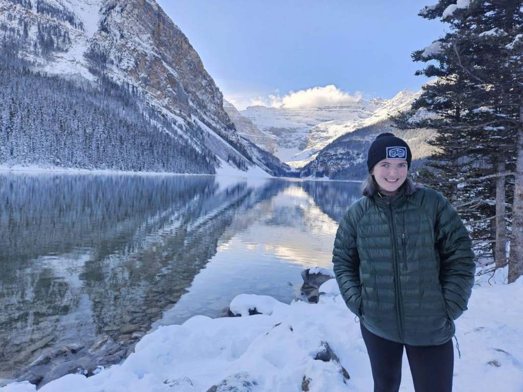 Abby wearing a winter hat and coat, smiling with a lake and snow covered mountains in the background.