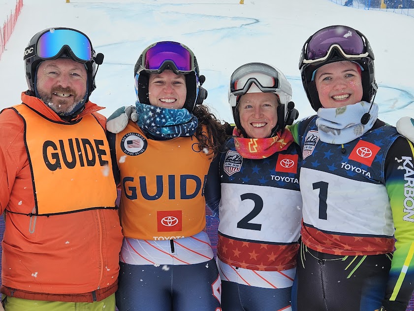 Abby, Chris, and two friends, a skier and guide who they completed against standing side by side. Everyone is smiling and wearing their ski suits and gear.  
