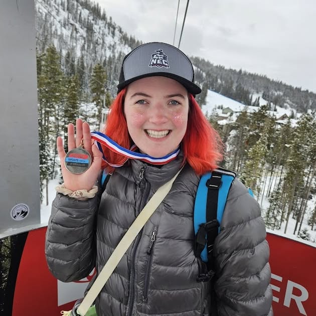 Abby smiling, wearing a gray winter coat and NEC baseball cap, holding up her medal that is around her neck. Snow covered mountains are in the background. 