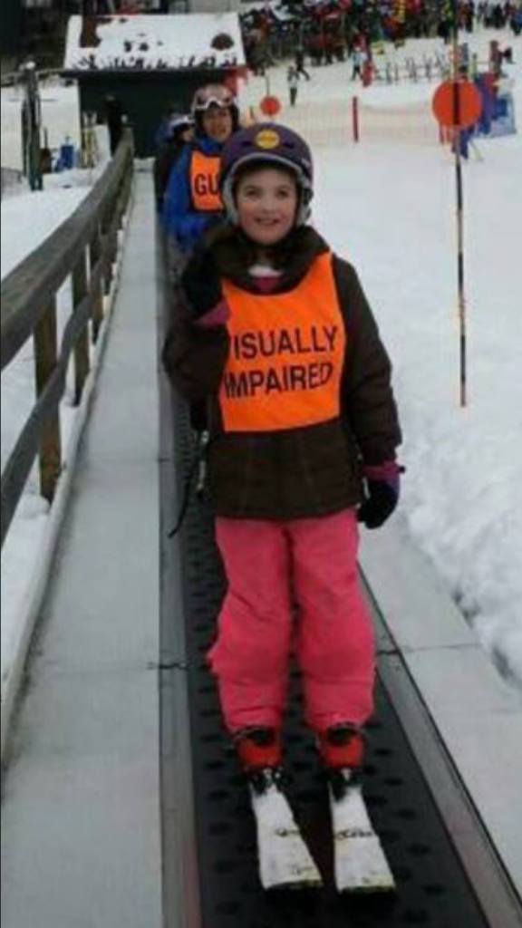 Young Abby, wearing skis, a helmet, orange snowpants and a ski bib with the text " visually impaired"  riding on the magic carpet. 