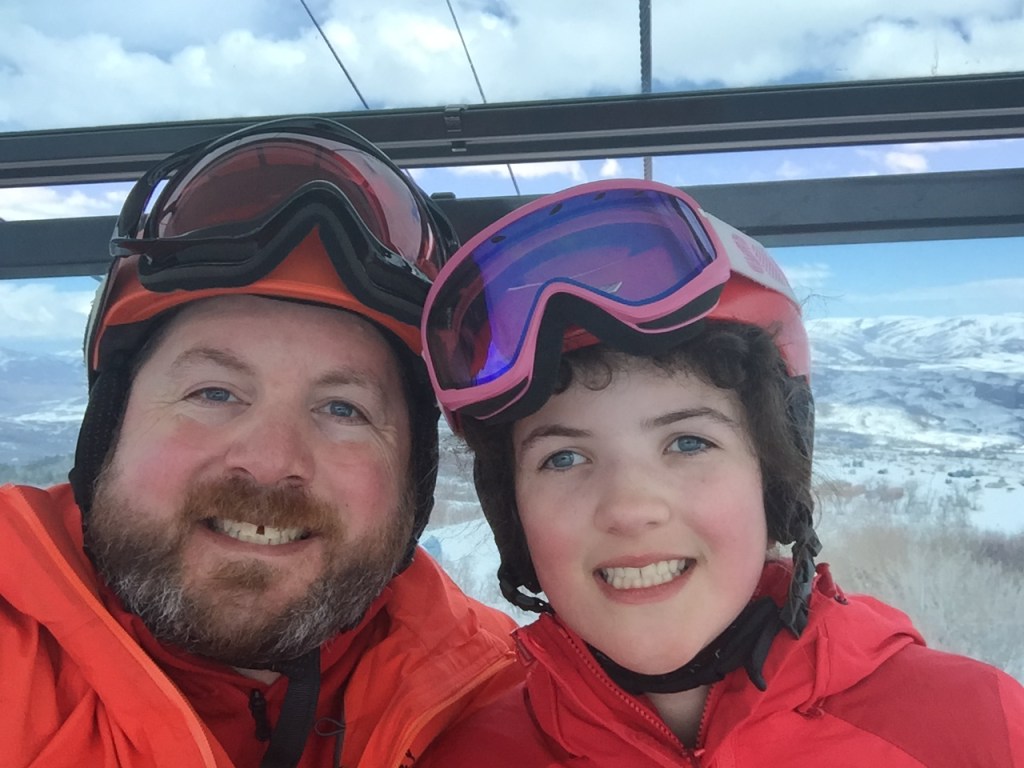 Young Abby and Chris, wearing matching orange ski suits and helmets smiling with snow covered mountains in background 